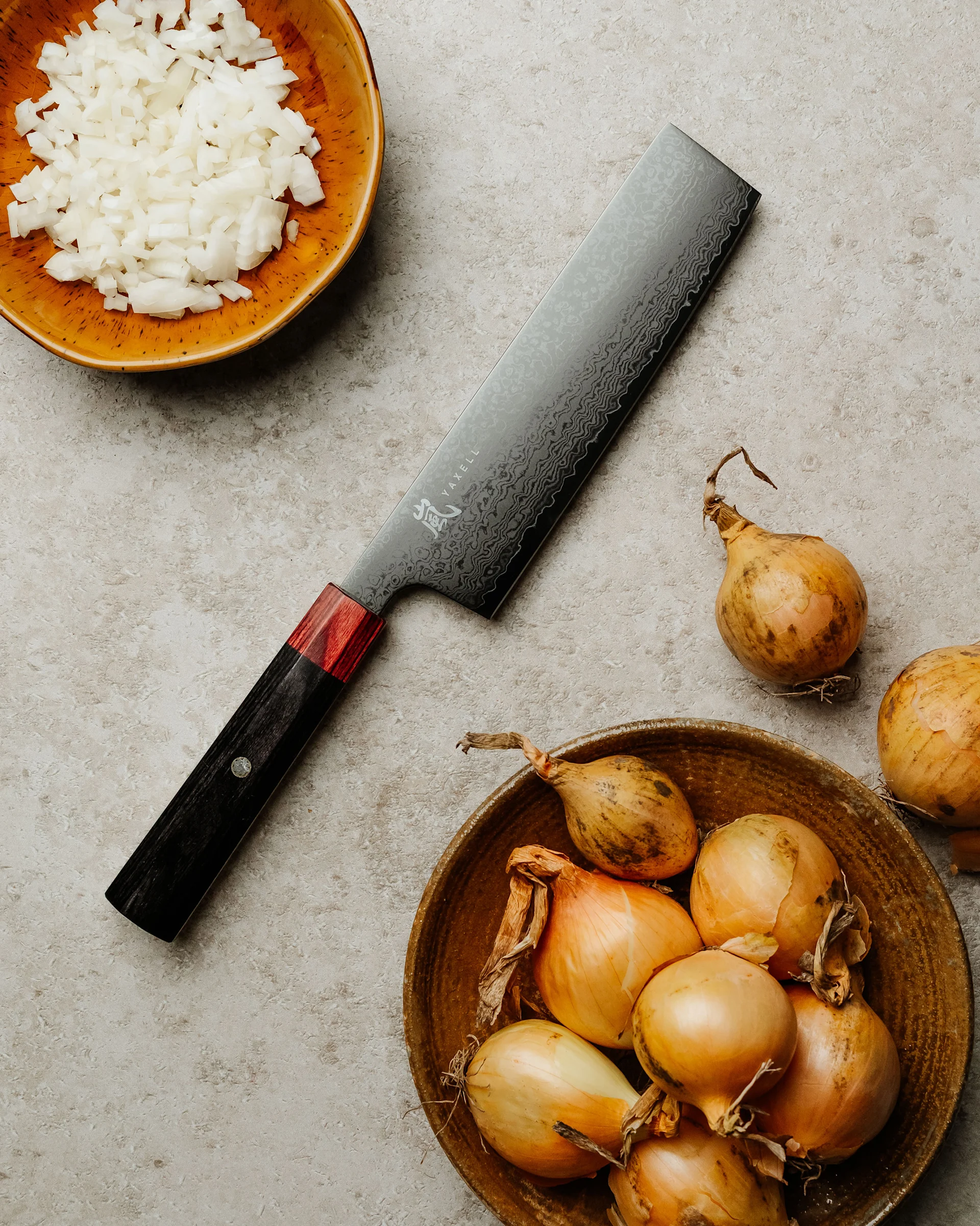 En kniv med mönstrat blad och rött/svart handtag ligger diagonalt. En skål med hackad lök och en med hela gula lökar bredvid.