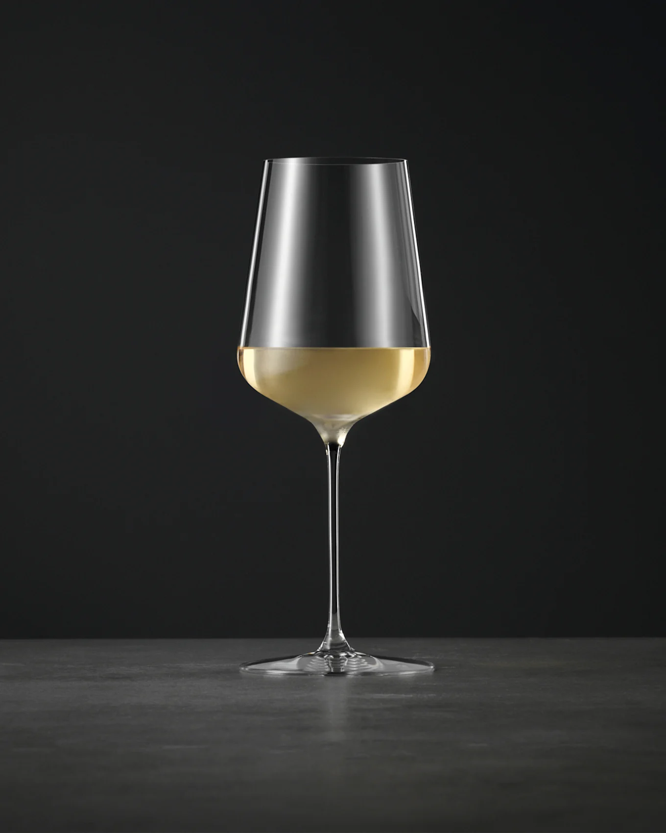Ett enkelt, elegant vinglas fyllt med vitt vin, på en mörk yta mot en svart bakgrund.