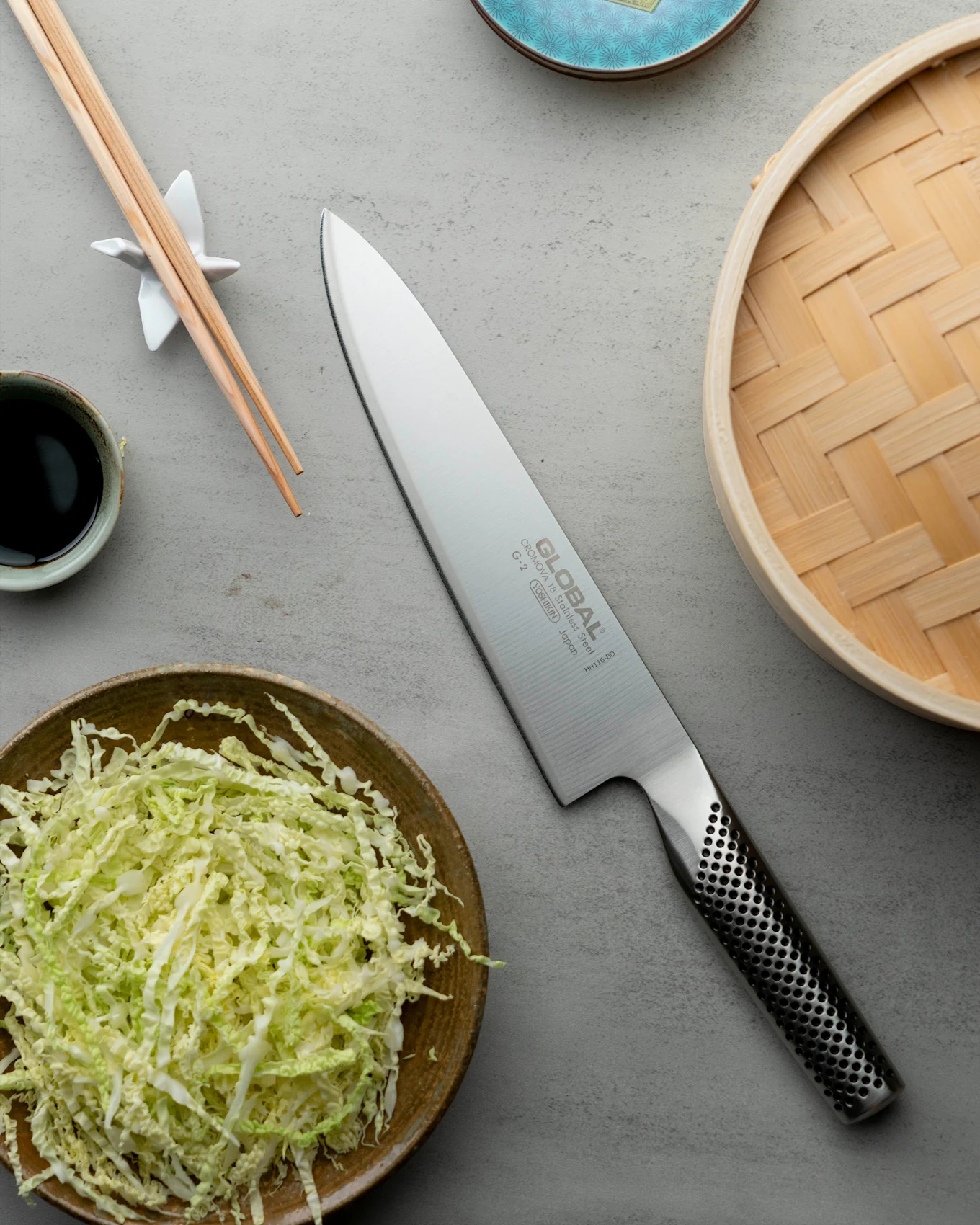 En Global G-2 kockkniv, strimlad kål, ätpinnar, sojasås och en bambuångare på en grå yta.