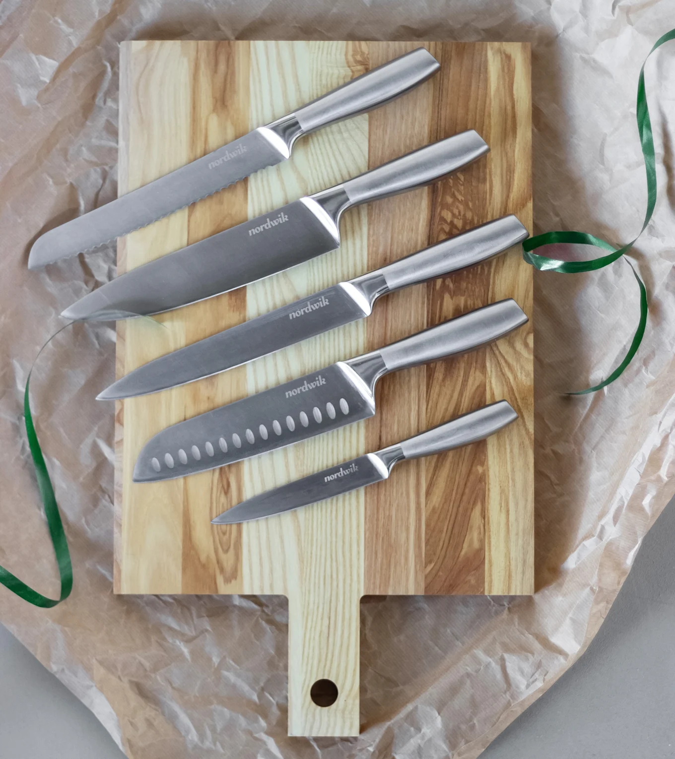 Ett set med fem Nordwik köksknivar i rostfritt stål, varierande i storlek, arrangerade på en ljus skärbräda i trä med ett grönt band.