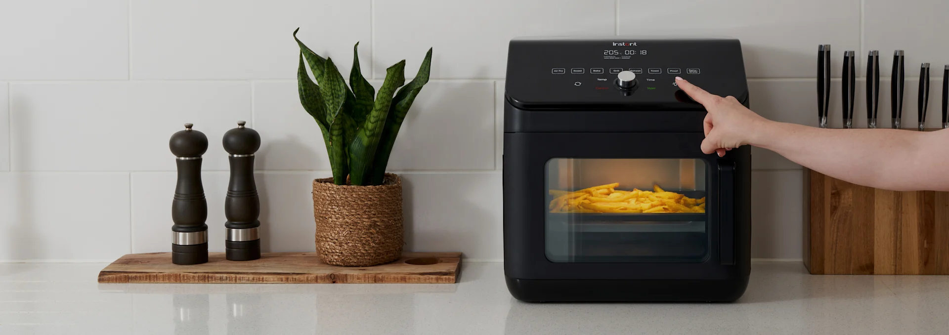 Hand trycker på knapp på svart airfryer med pommes frites, bredvid kryddkvarnar, en växt och knivblock på köksbänken.
