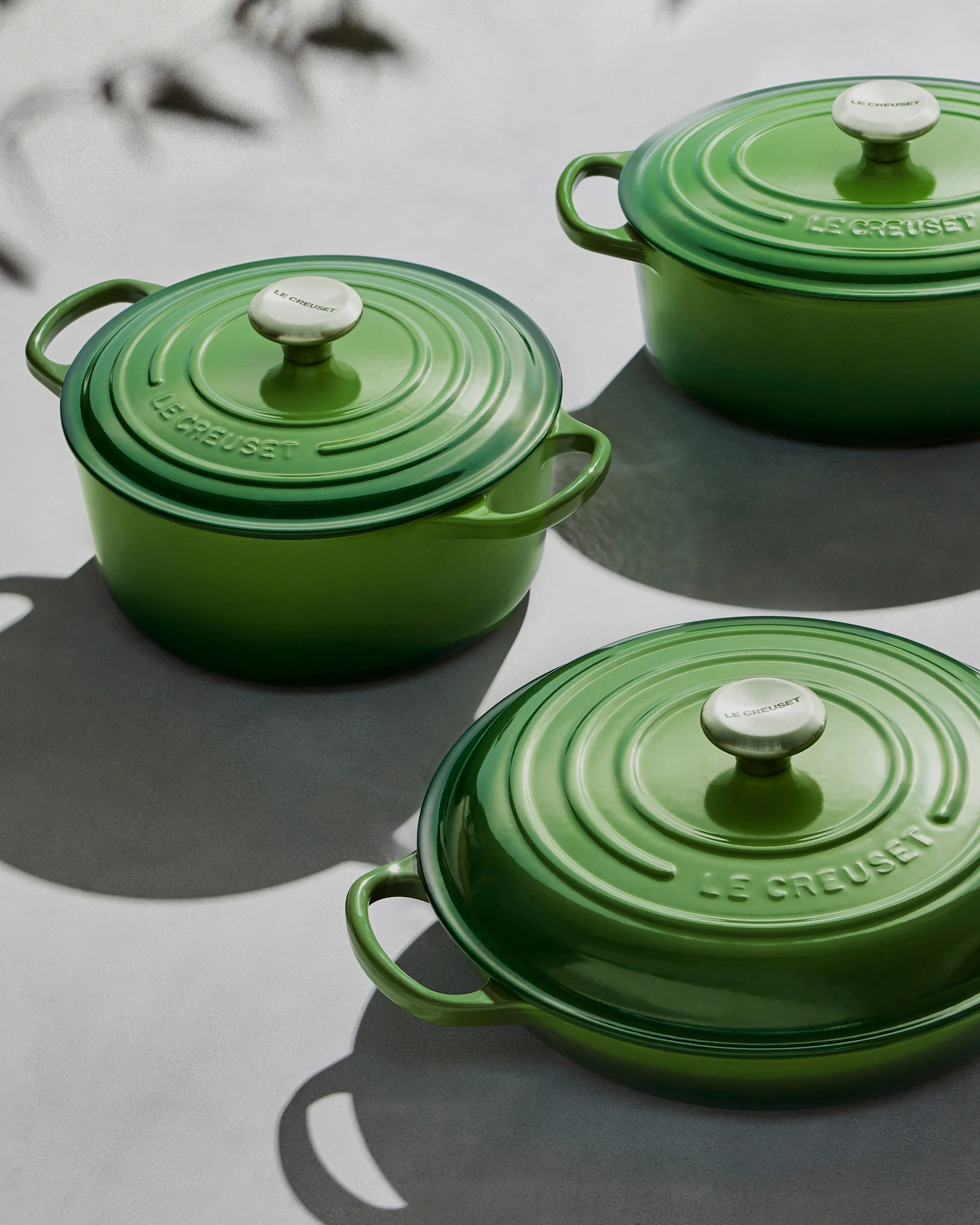 Tre gröna Le Creuset grytor med lock och silverknoppar på en ljus yta med skuggor.