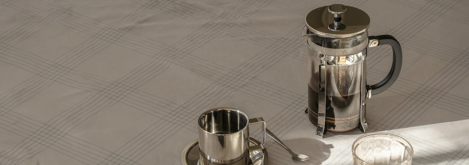Kaffepress med kaffe, en metallkopp och fat med sked på en ljus, rutig duk i solljus.