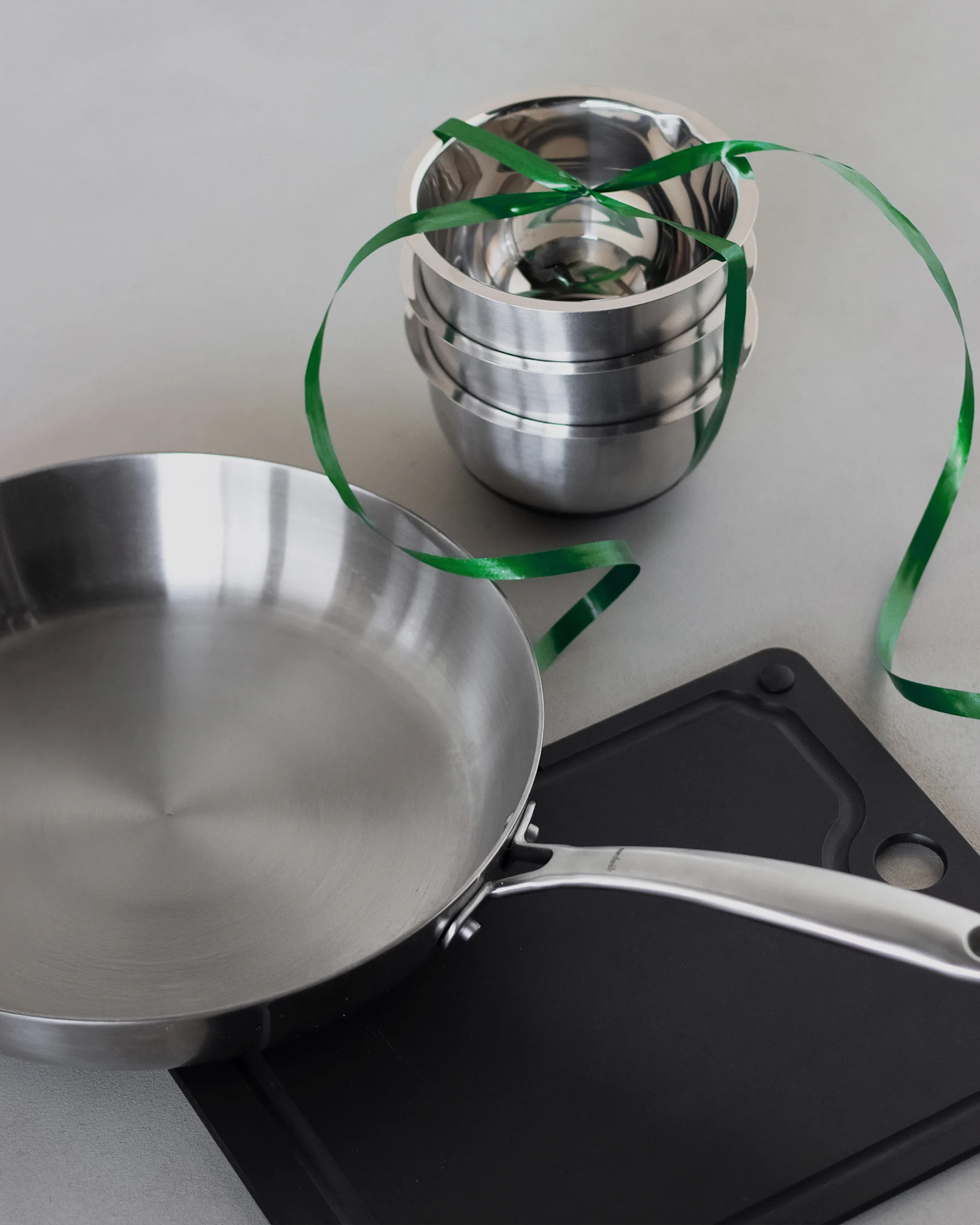 En stekpanna i rostfritt stål, en stapel metallskålar bundna med ett grönt band och en svart skärbräda.