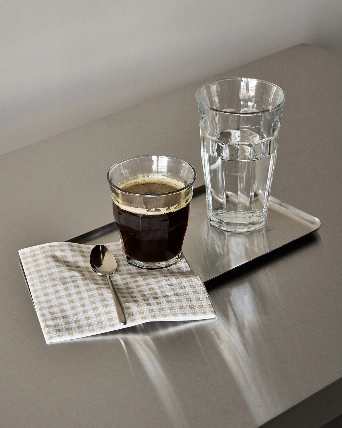 Glas svart kaffe, glas vatten och en sked på en rutig servett på en metallbricka.